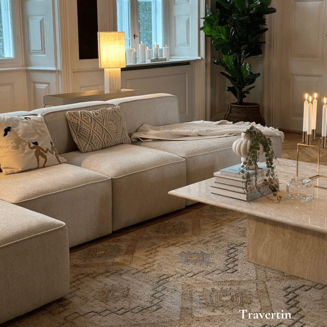 Elegant stue med beige sofa, travertin sofabord, planter og lys, der skaber en atmosfærisk indretning.